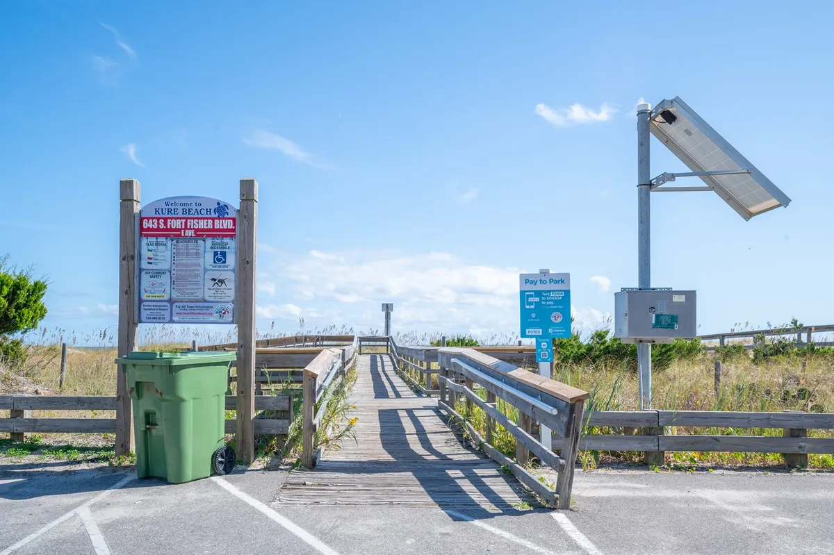 A boardwalk leading to the beach, marked by a beach access sign and surrounded by dune grass. Subtle details like a solar panel and pay-to-park sign highlight the functionality and welcoming access to the shoreline.