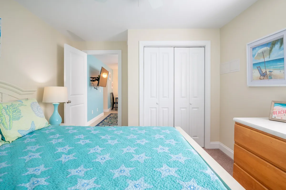 A tranquil bedroom with a bed covered in a star-patterned quilt, a nightstand with a lamp, a dressing area with a dresser, and cheerful artwork. The space is bright, calm, and perfect for restful nights or quiet mornings.