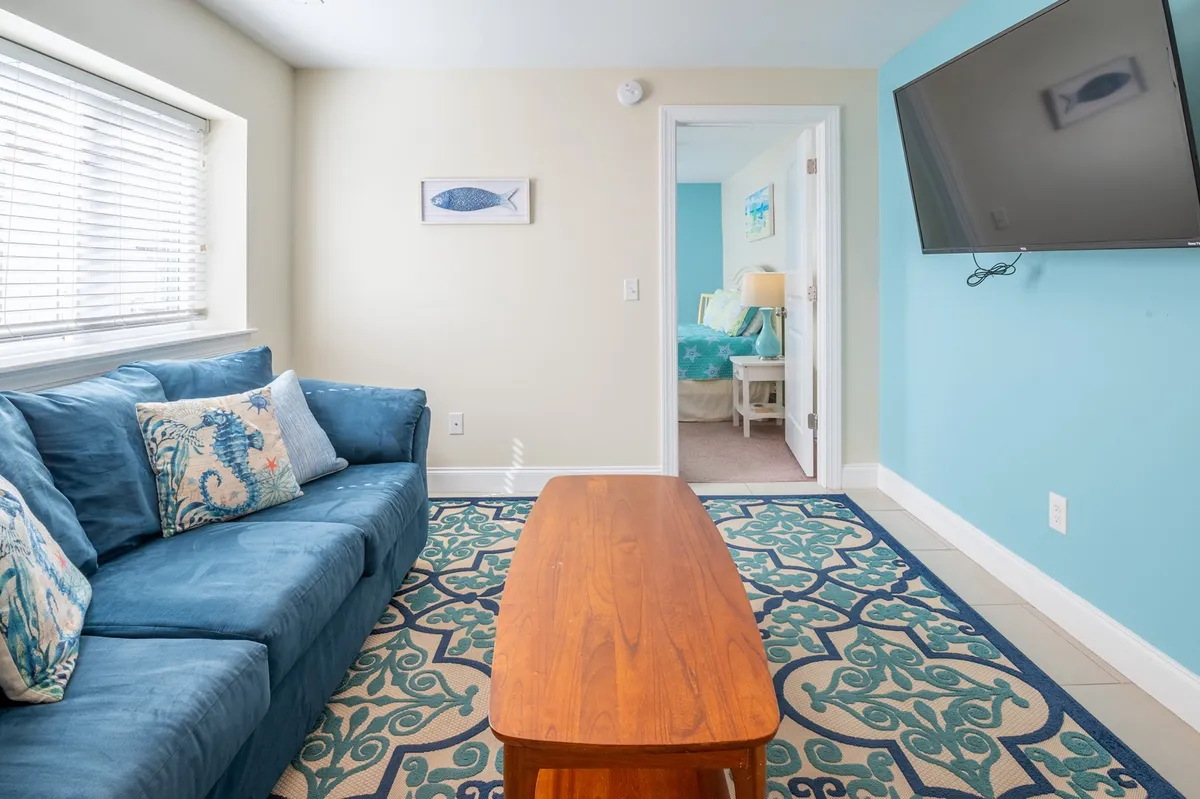 A comfortable living area featuring a blue sofa with decorative pillows, a wooden coffee table, a textured rug, wall art with a fish motif, and a television mounted on the wall. The space feels relaxed and is designed for both socializing and quiet downtime.