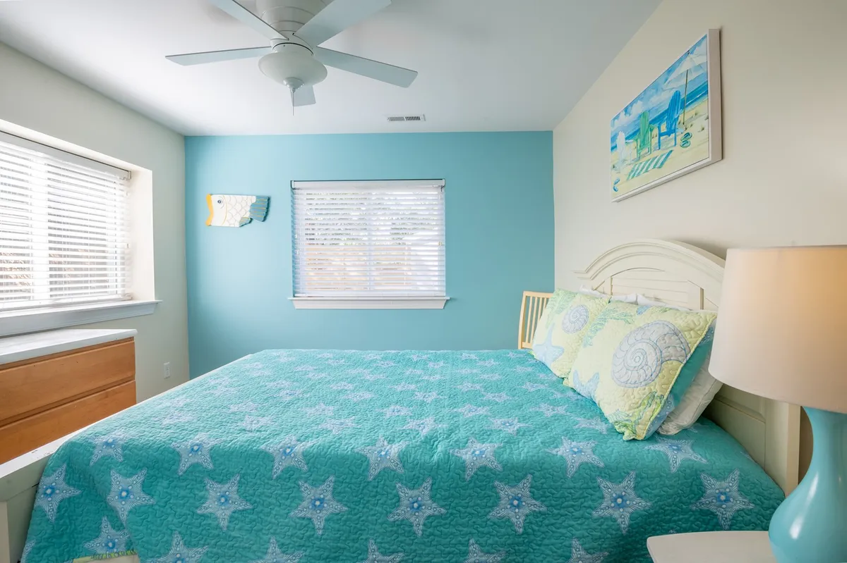 This calming bedroom displays a bed with a star-patterned quilt, decorative wall art, a window with blinds, and a nightstand with a lamp. The gentle natural light and soft décor create an inviting space ideal for rest and privacy.