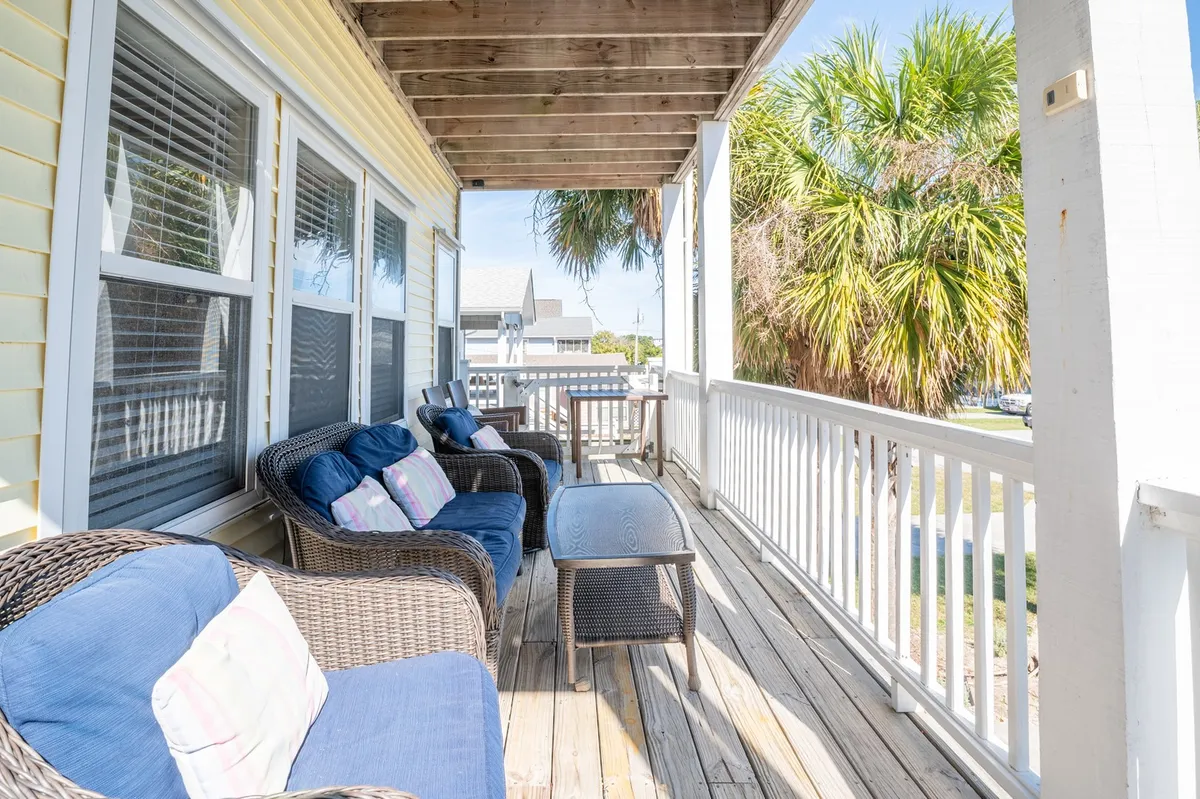 A welcoming front porch featuring wicker chairs with blue cushions and a wooden table, framed by palm trees in the background. The seating area is arranged to invite conversation and relaxation, perfect for enjoying the coastal breeze and outdoor living.