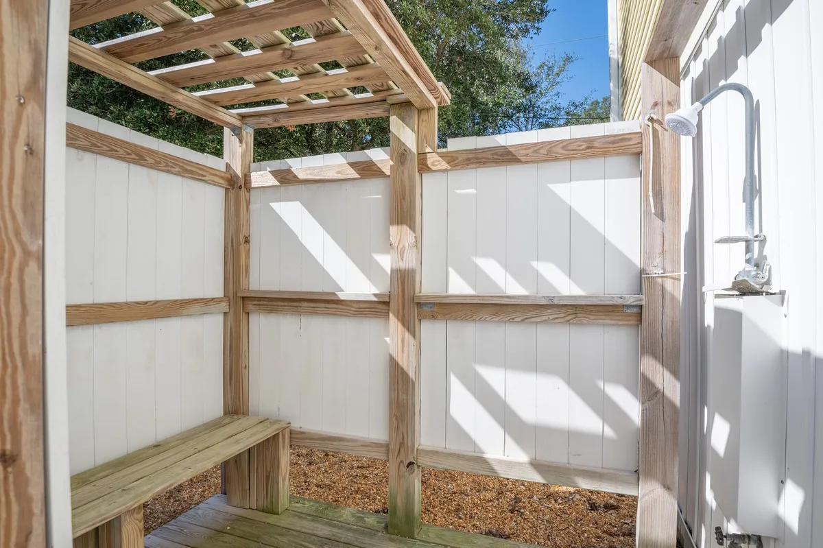 An outdoor shower area with a wooden bench and pergola structure, surrounded by greenery. The space offers privacy and a refreshing atmosphere, perfect for rinsing off after a day at the beach.