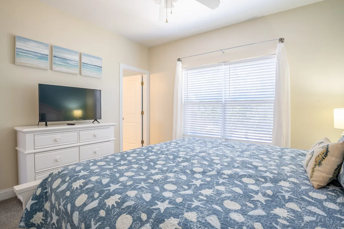 This image features a serene bedroom with a blue patterned quilt on the bed, a white dresser with a TV, and a large window covered by sheer curtains. The natural light streaming in enhances the calming, hotel-inspired vibe of the space, ideal for restful nights.