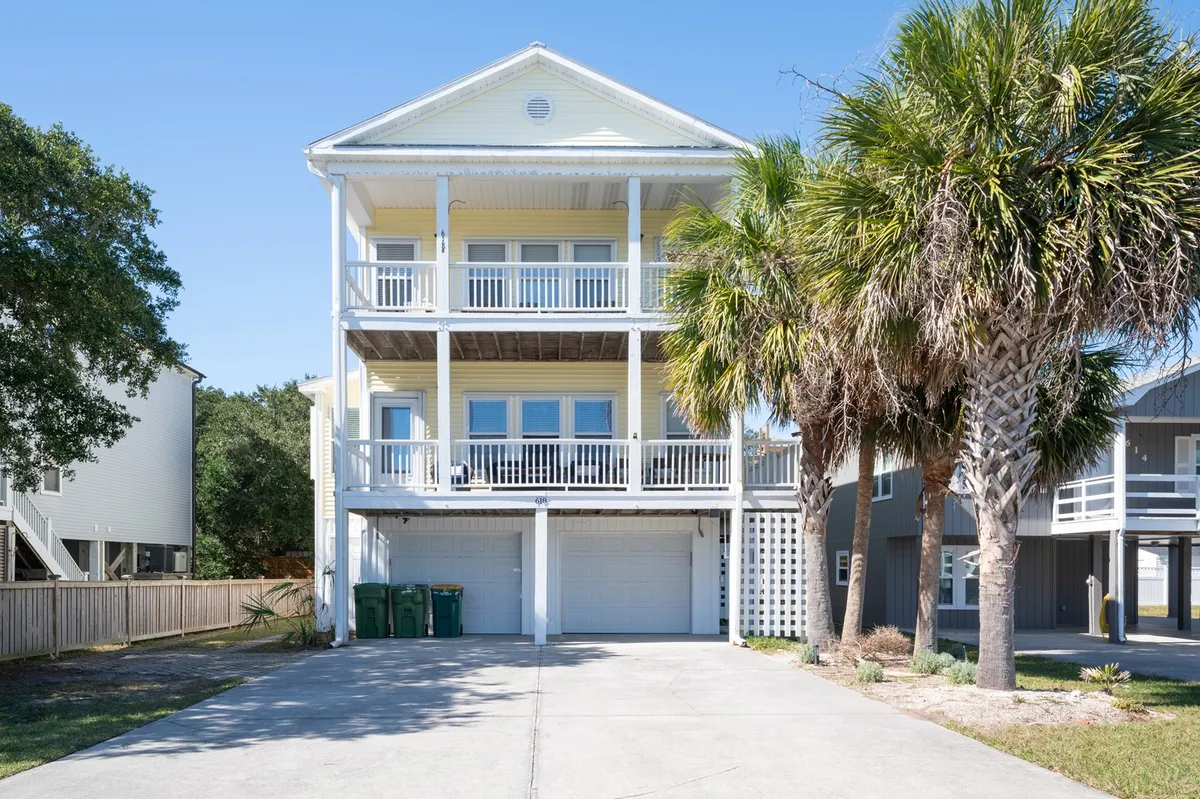 The image showcases the cheerful yellow exterior of the house with inviting balconies surrounded by palm trees. The setting suggests a relaxed, coastal atmosphere perfect for gathering outdoors, and hints at the home's proximity to local attractions and the beach.