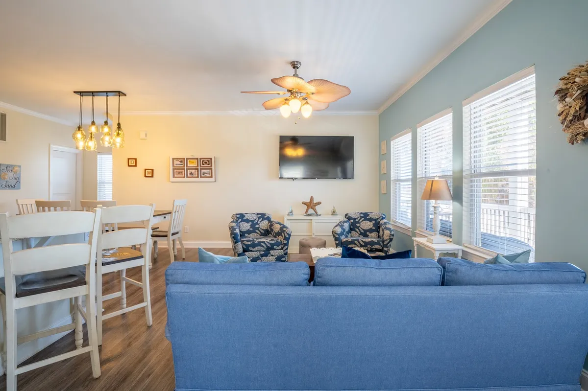 This image depicts a bright, inviting living and dining area, featuring a blue sofa, floral chairs, a dining area, and a TV, with a hanging light fixture above. The space is open and airy, suggesting a spot for gathering after a day out, with a glimpse of sunlight hinting at an easy transition between indoor comfort and outdoor adventure.