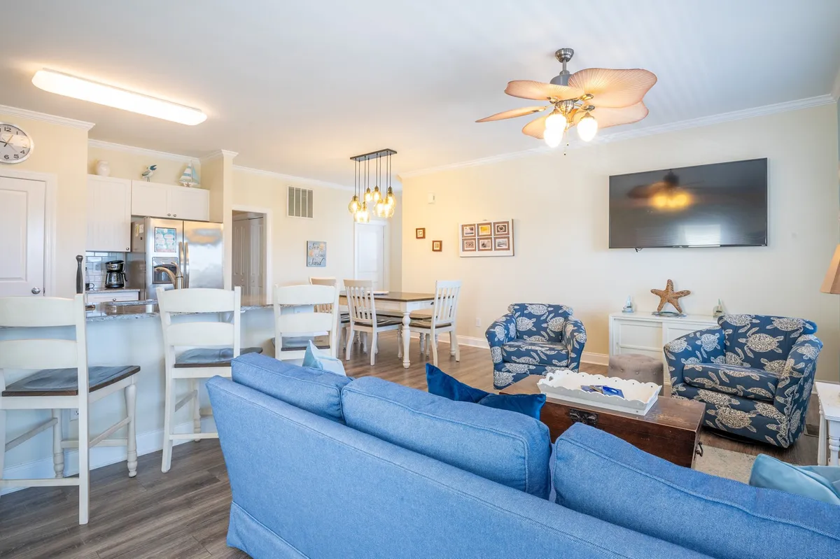 A bright, open-concept living and dining area with a blue sofa, spacious dining table, kitchen counter, and decorative wall art. The layout encourages group interaction, with ample seating for family meals, games, or movie nights after a day of adventure.