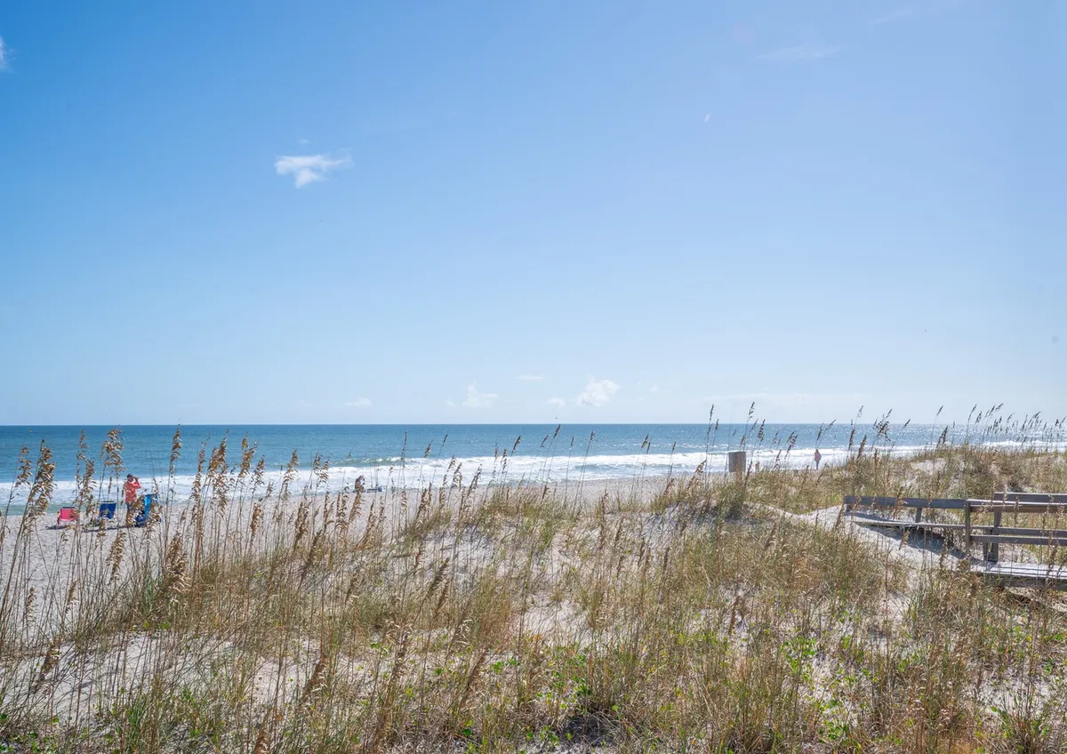A classic beach scene with a sandy shoreline, ocean waves, dune grass, chairs, and a boardwalk. The environment is tranquil and inviting, encapsulating the spirit of coastal adventures and outdoor fun.